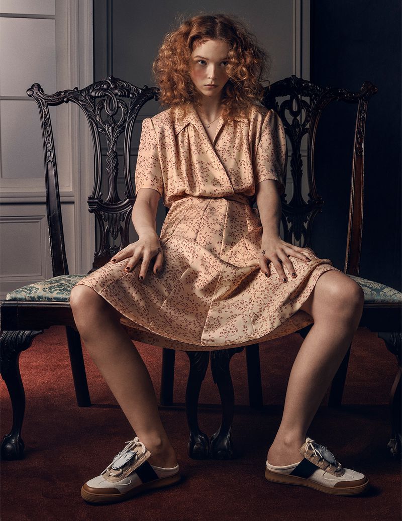 Elegant woman sits on a carved wooden chair wearing a peach-colored dress and shoes in a studio setting with soft lighting.