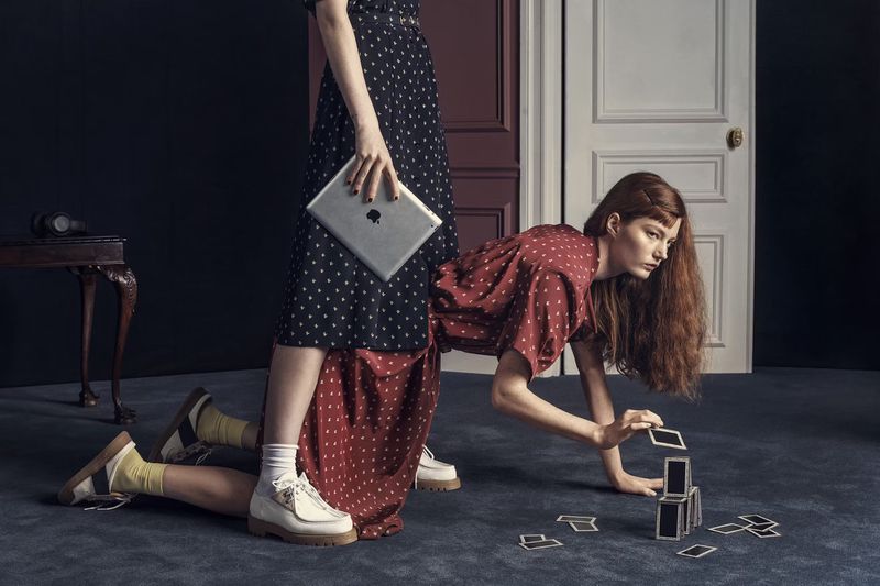 Two women pose indoors: one holds a tablet while standing, wearing a long polka-dot dress and the other kneels building a card tower.