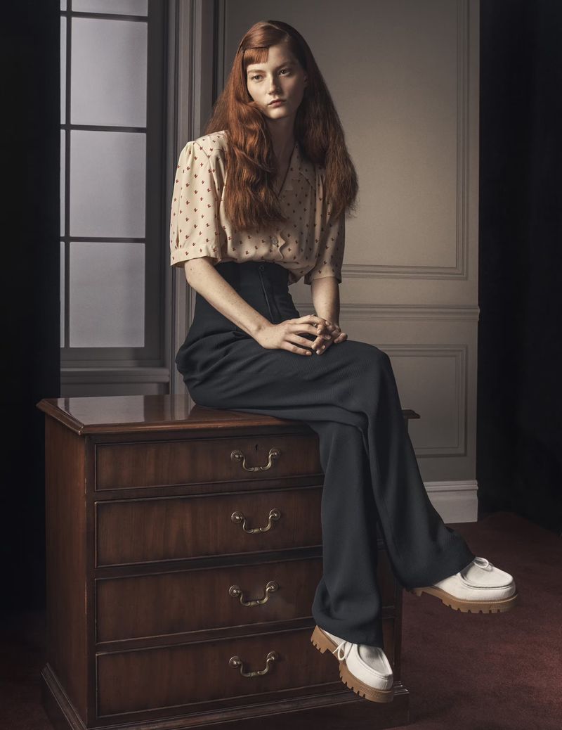 A woman with long red hair sits on a dark wood dresser in a photo studio. She wears a vintage-style blouse and high-waisted trousers.