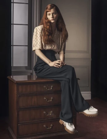 A woman with long red hair sits on a dark wood dresser in a photo studio. She wears a vintage-style blouse and high-waisted trousers.