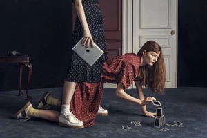 Two models in patterned dresses posed together indoors with a tablet and cards on a carpeted floor for W Magazine.