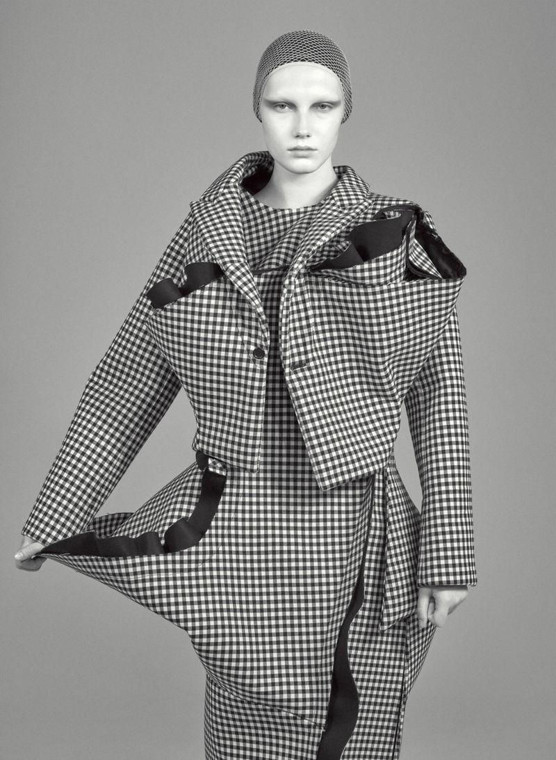 A fashion model presents a black and white houndstooth jacket and wrap skirt with black trim against a solid gray backdrop in a studio shot.