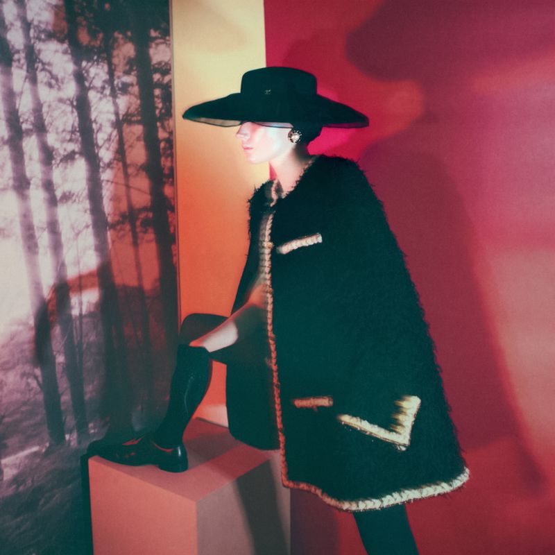 Fashion shot of a female model in a black coat and hat, kneeling on a block in front of a forest scene with red and orange lighting.