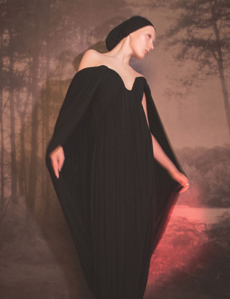 Fashion shoot of a slender woman wearing a black off-the-shoulder dress in front of a muted forest backdrop, bathed in soft light.
