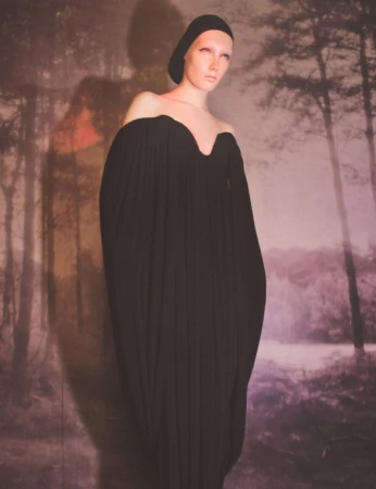 An adult female model with pale skin wears an off-the-shoulder black dress in front of a backdrop with a blurry forest and lake.