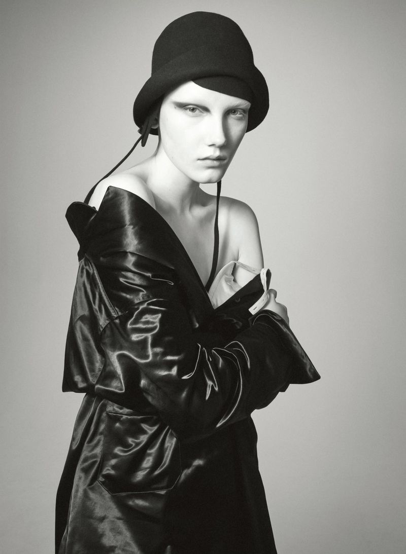Woman in black hat and shiny coat poses in studio with neutral background. High-fashion, minimalist composition, dramatic lighting.