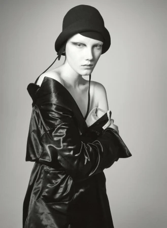 Woman in black hat and shiny coat poses in studio with neutral background. High-fashion, minimalist composition, dramatic lighting.