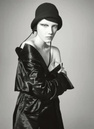 Model wearing a black cloche hat and dark satin coat against a plain neutral background for W Magazine.