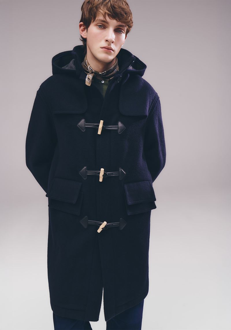 Adult male poses indoors wearing a navy duffle coat with toggle closures, a scarf, and blue jeans, set against a gradient gray background.