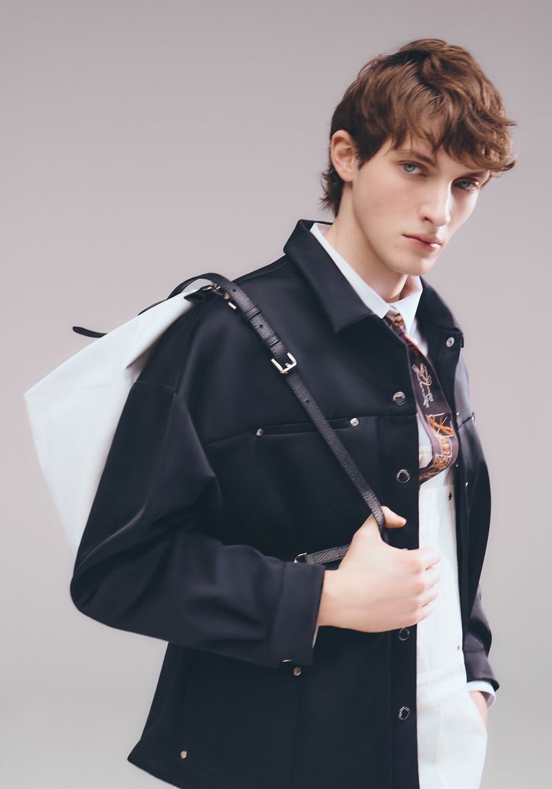 A stylish adult male in a dark jacket and white shirt, carries a white tote bag, stands against a muted background in a fashion-forward pose.
