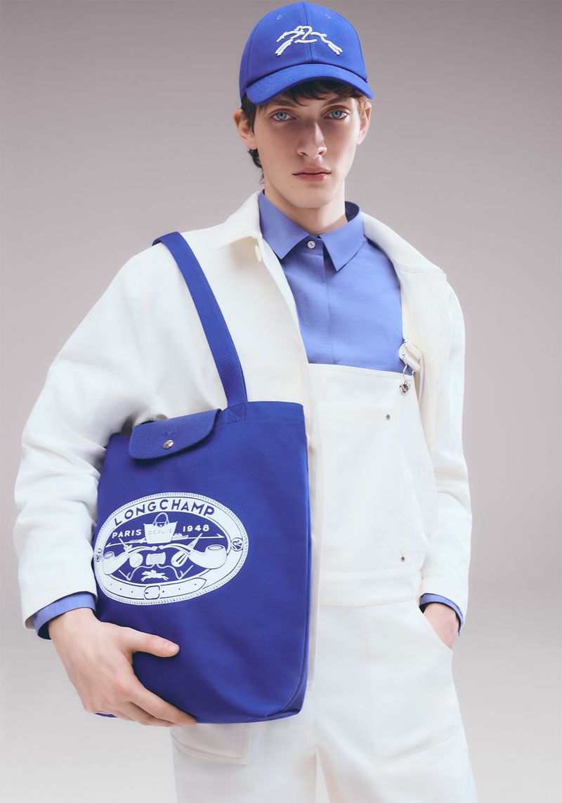 Male fashion model poses with Longchamp tote bag, wearing matching blue baseball cap and shirt with a white workwear jacket and pants.
