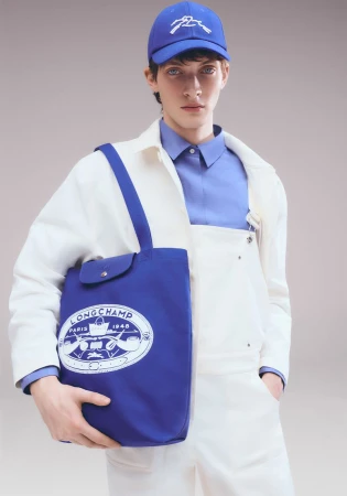 Male fashion model poses with Longchamp tote bag, wearing matching blue baseball cap and shirt with a white workwear jacket and pants.