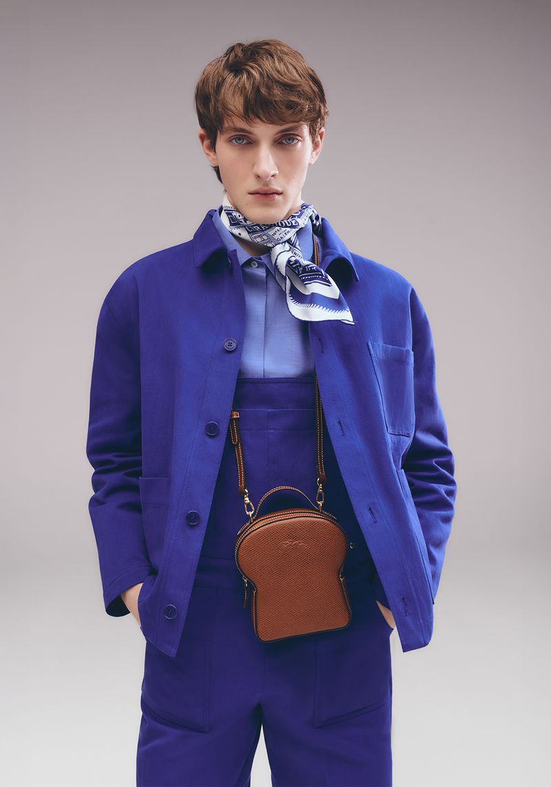 A fair-skinned male model wears a blue suit and a brown, toast-shaped handbag against a neutral backdrop, showcasing a unique fashion statement.