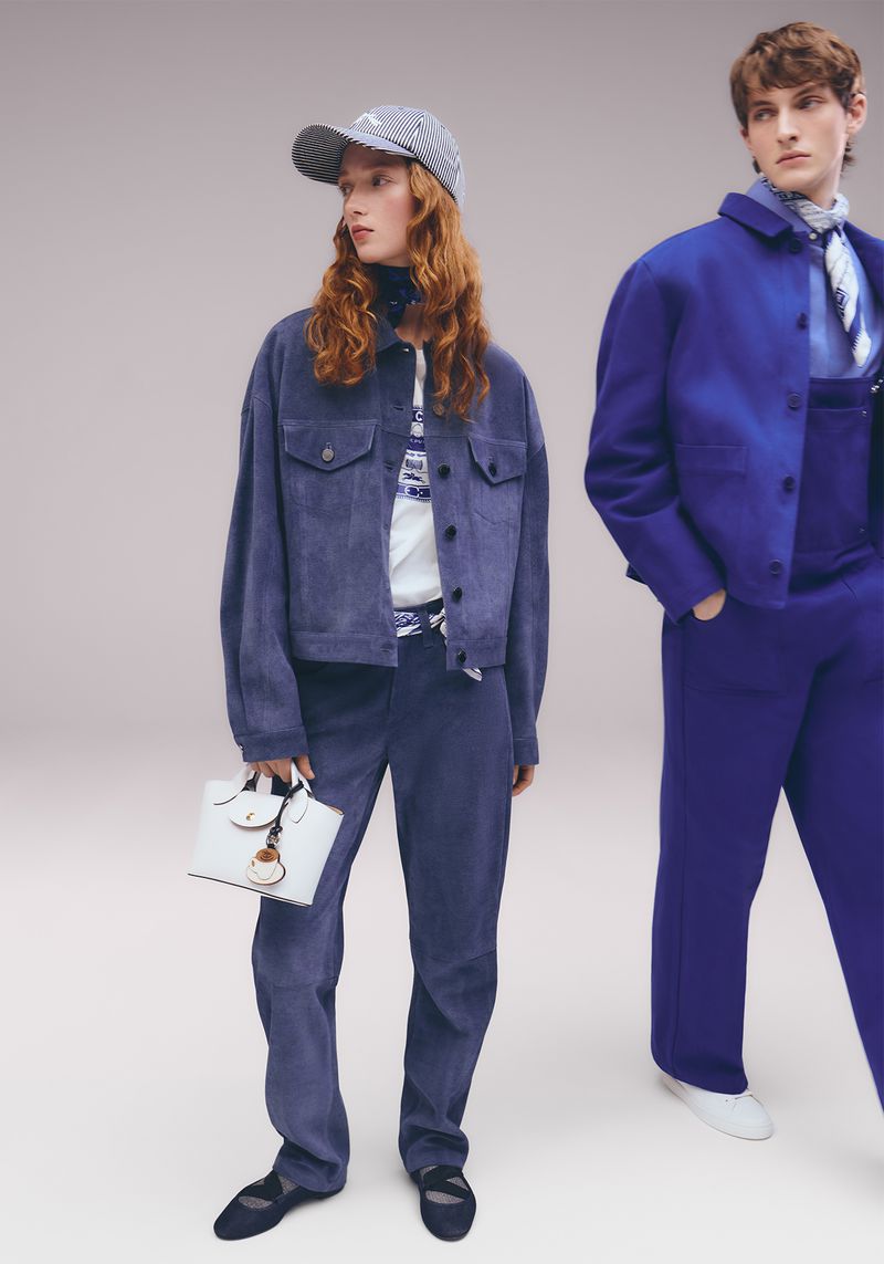 Two people model blue outfits and accessories in a studio setting, with one person holding a white handbag, showcased in a fashion editorial.