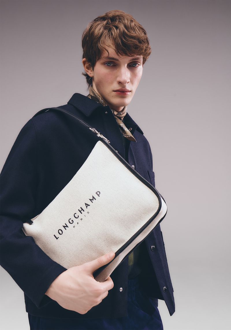Fashion shot of a male model holding a Longchamp handbag, wearing a navy jacket and patterned scarf, against a grey backdrop.