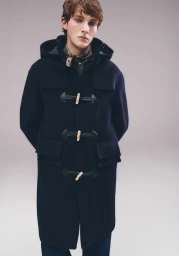 Model wearing a dark hooded duffle coat with toggle closures against a plain light grey studio background.