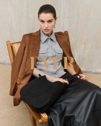 Model seated in a wooden chair wearing a grey buttoned polo shirt, brown jacket, and black skirt holding a black handbag.