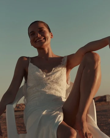 A cheerful young woman in a white dress sits on a chair outdoors, smiling against a clear sky and arid landscape backdrop.