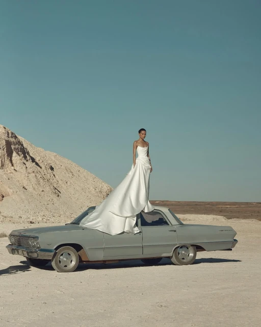 Woman in white strapless gown standing on the hood of a gray vintage car with desert and pale blue sky in the background.