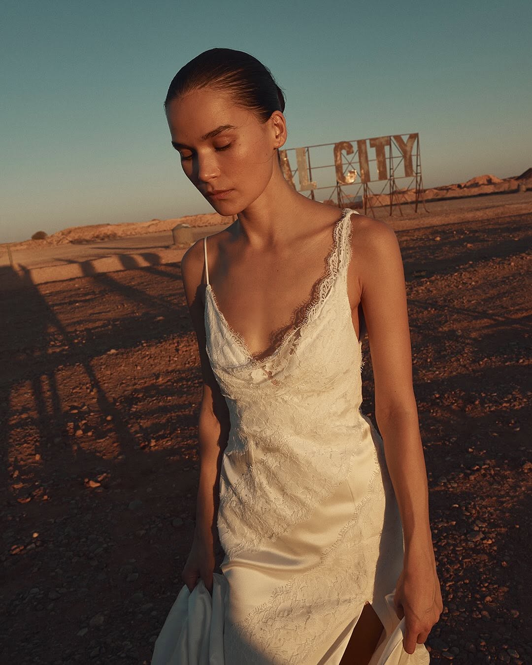 Woman in a lace gown in a desert setting with blue skies and a vintage sign in the background; warm, natural lighting.