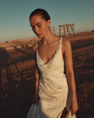 Woman in a lace gown in a desert setting with blue skies and a vintage sign in the background; warm, natural lighting.