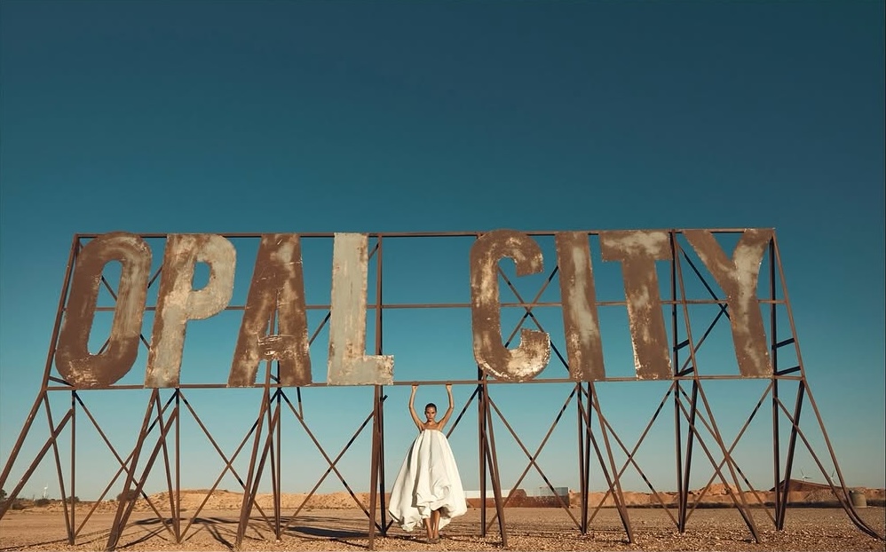 Woman poses in front of an "Opal City" sign in an outdoor desert landscape, under a clear blue sky, in a fashion editorial shot.