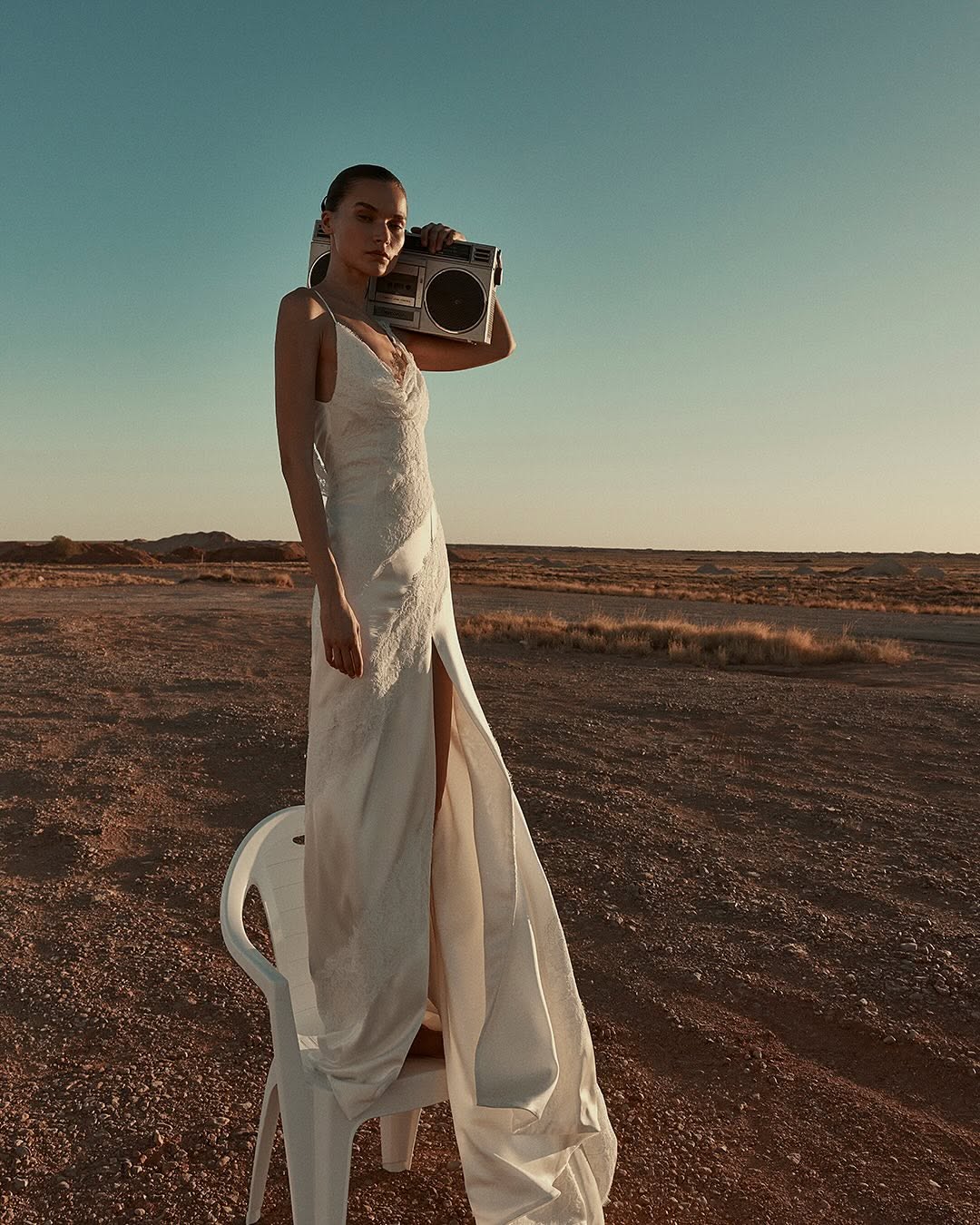 Woman in a white dress stands on a plastic chair in a desert landscape, holding a boombox. Clear sky, medium shot, fashion photography.