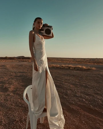 Woman in a white dress stands on a plastic chair in a desert landscape, holding a boombox. Clear sky, medium shot, fashion photography.