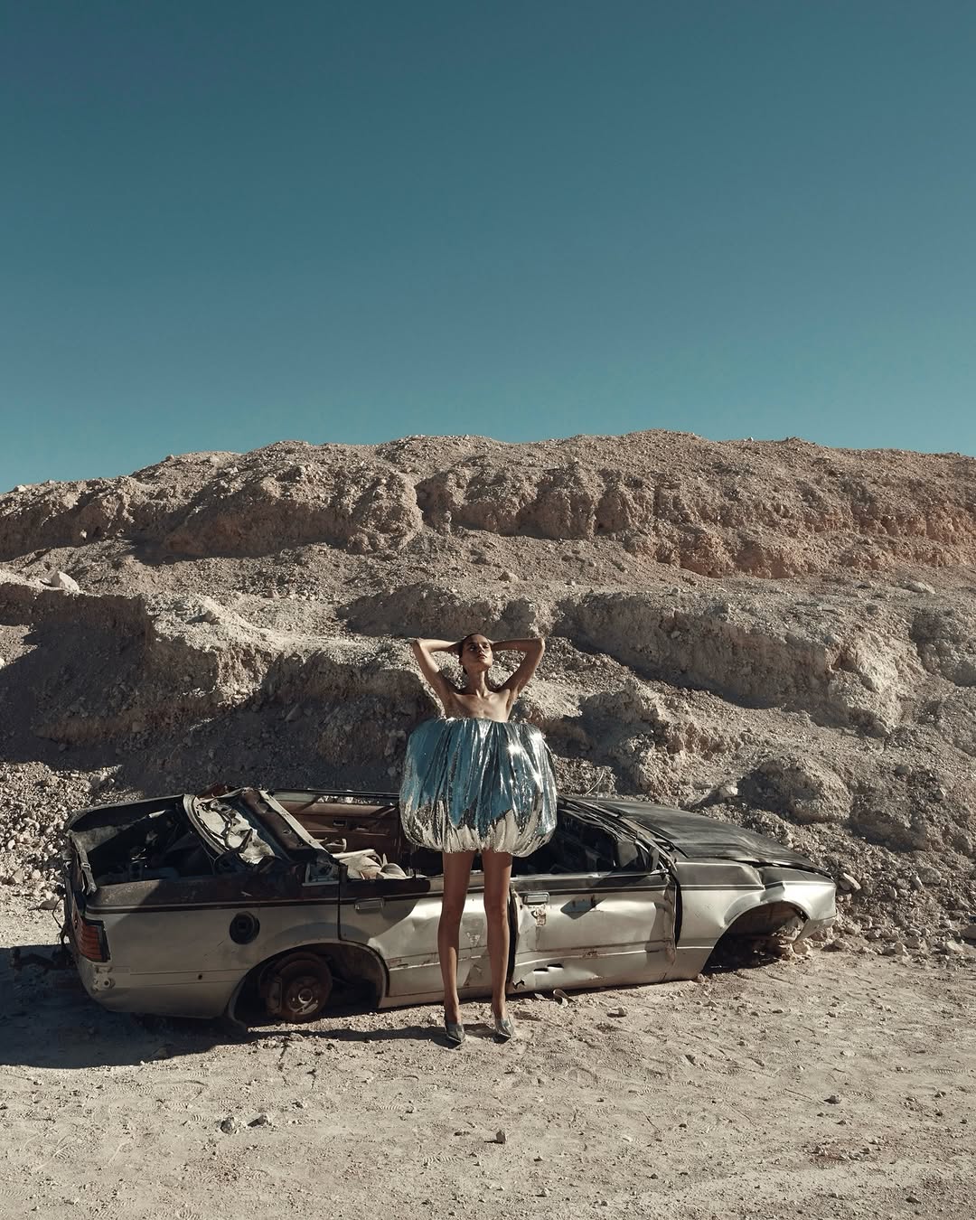 Fashion shot of a slender woman in a reflective dress posing next to a wrecked car against a desert landscape with a clear blue sky.