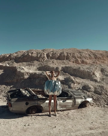 Fashion shot of a slender woman in a reflective dress posing next to a wrecked car against a desert landscape with a clear blue sky.