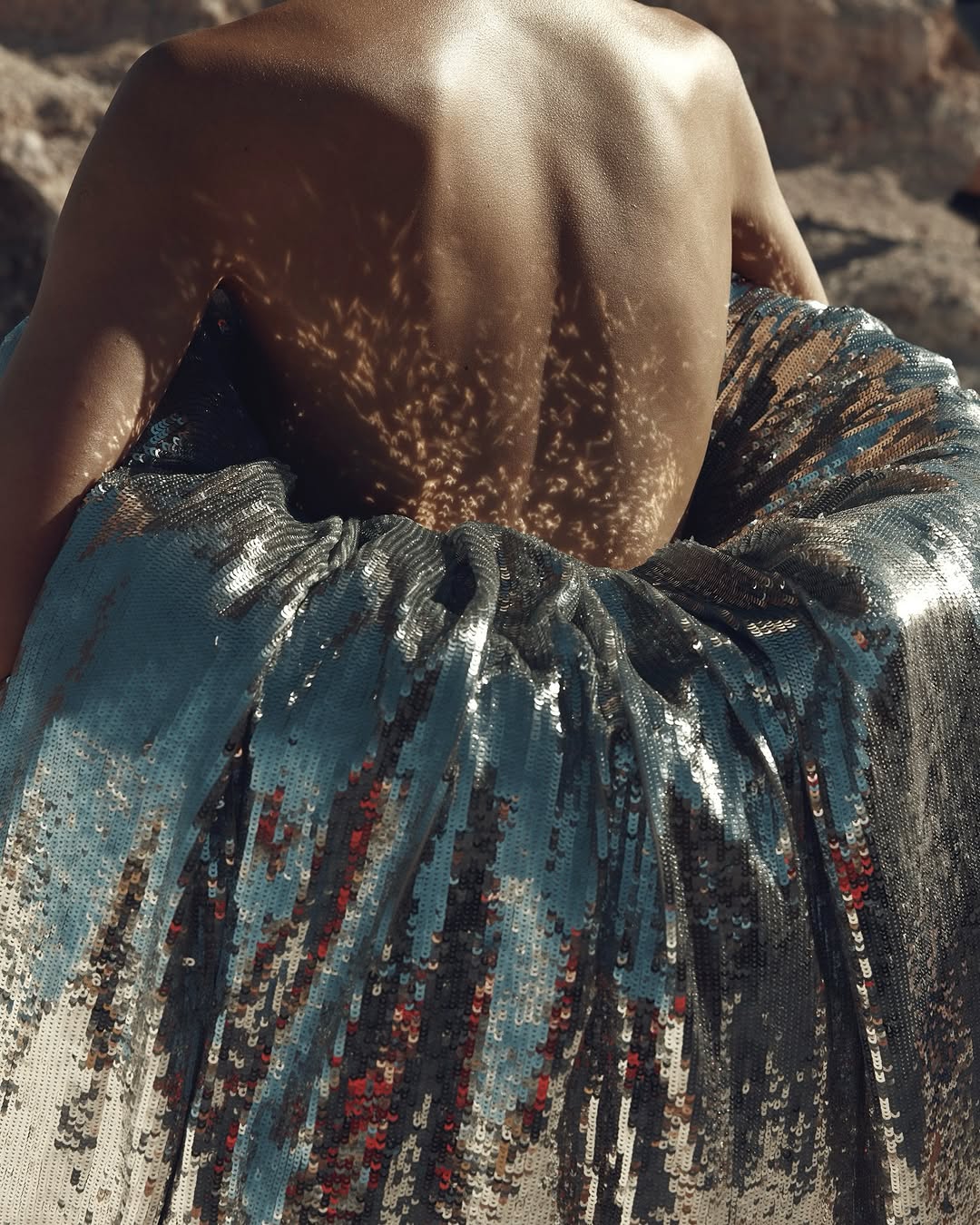 A back view of an adult female with shadows on skin, wearing a shiny silver skirt, posing outdoors in soft, diffused sunlight.