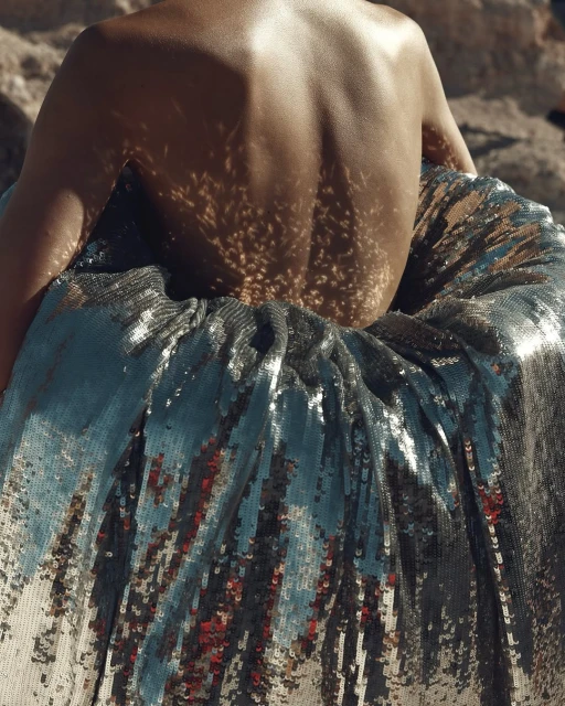 A back view of an adult female with shadows on skin, wearing a shiny silver skirt, posing outdoors in soft, diffused sunlight.