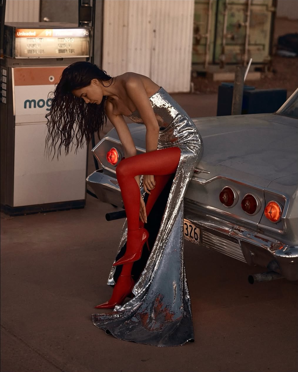 Woman in sequin dress posing on vintage car in desert setting near gas station. Red stockings and heels, evening lighting.
