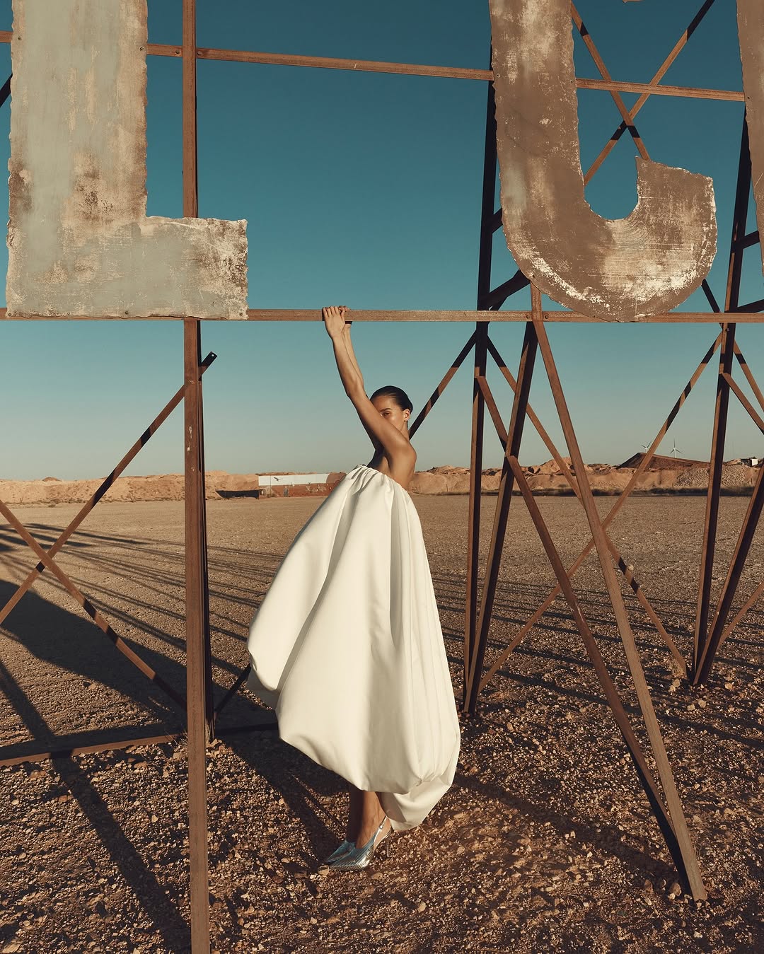 Woman in voluminous white skirt poses holding the frame of a giant metal sign in a desert under a clear blue sky.