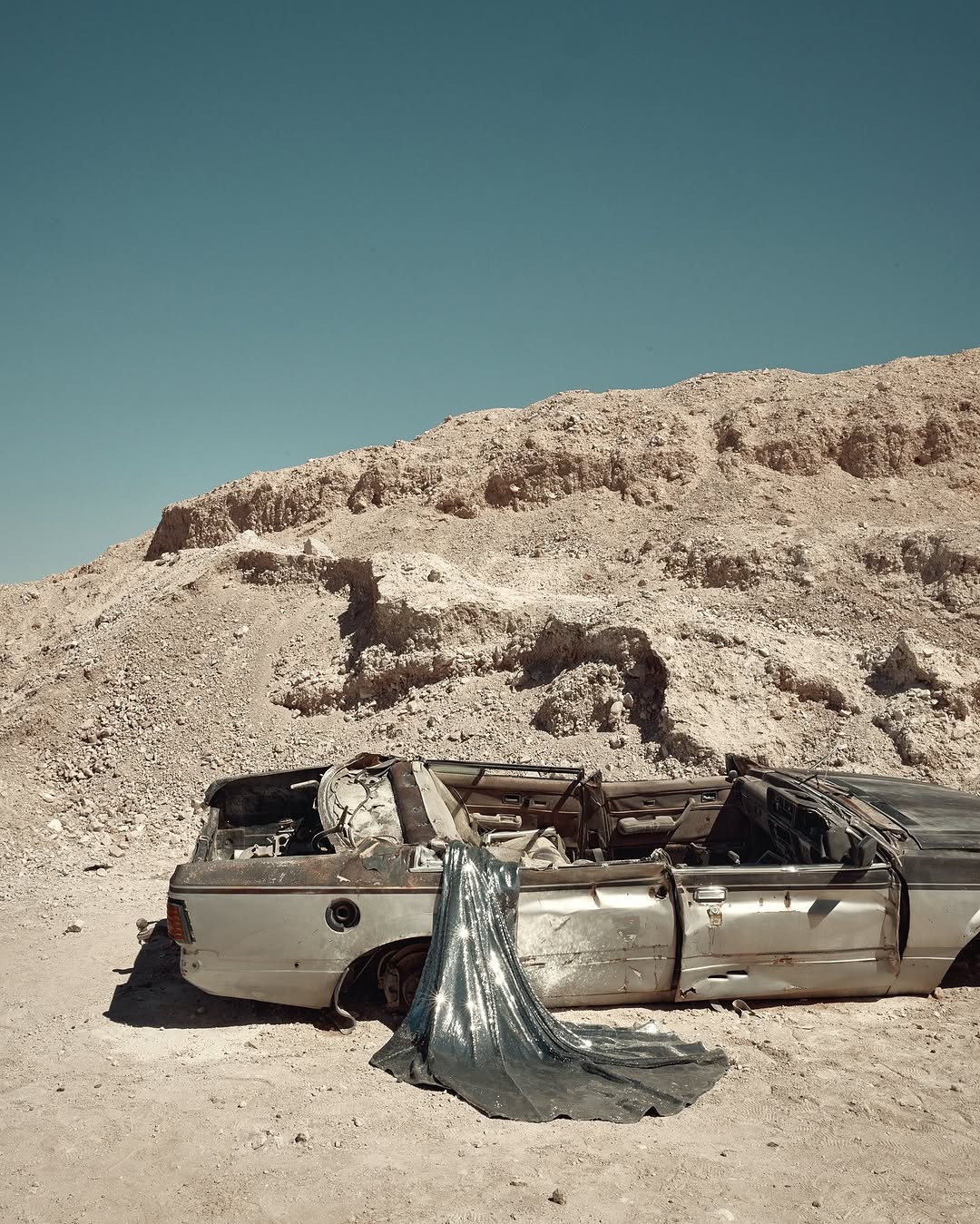 Wrecked car in desert landscape with flowing silver fabric, set against a backdrop of sun-baked earth and clear blue skies.