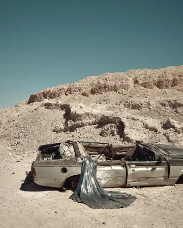 Wrecked car in desert landscape with flowing silver fabric, set against a backdrop of sun-baked earth and clear blue skies.