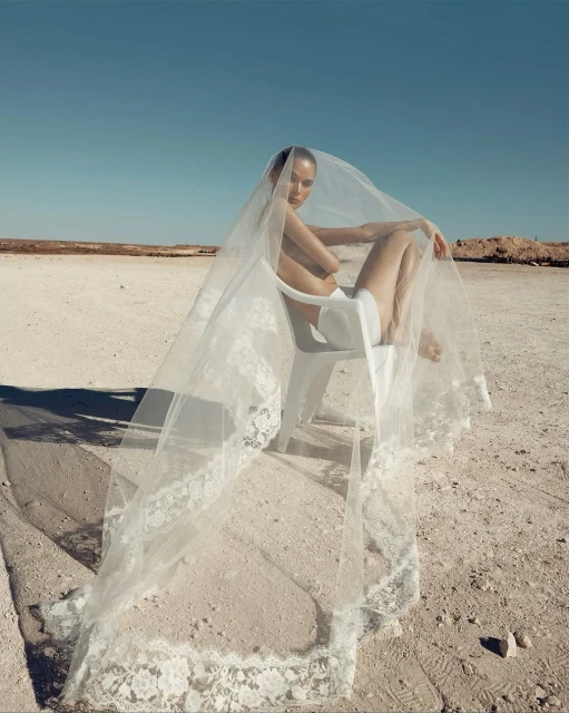 Woman in a veil sits on a chair in a desolate landscape. The bright blue sky contrasts with the beige and brown earth tones.