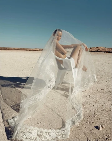 Woman in a veil sits on a chair in a desolate landscape. The bright blue sky contrasts with the beige and brown earth tones.