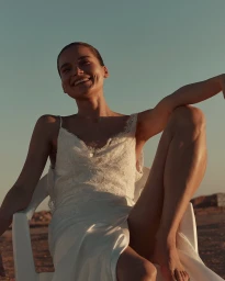 Smiling woman in a white slip dress sitting on a white chair outdoors against a clear sky for KYHA Bride Moon Plain Collection.