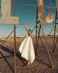 Model in voluminous white gown hanging from large rusty metal letter frame outdoors in the desert