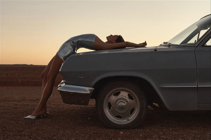 Model in a silver sequin minidress arching over the hood of a vintage car at sunset in a desert landscape