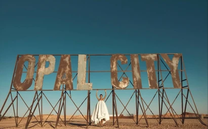 Model in a voluminous white bridal gown standing beneath a large rusted Opal City sign in a desert landscape under a clear sky.