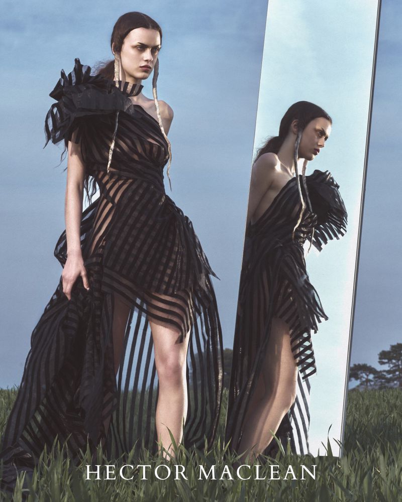 A fashion shot of a model in a long black striped dress with reflection in a standing mirror in a grassy field under a blue sky.