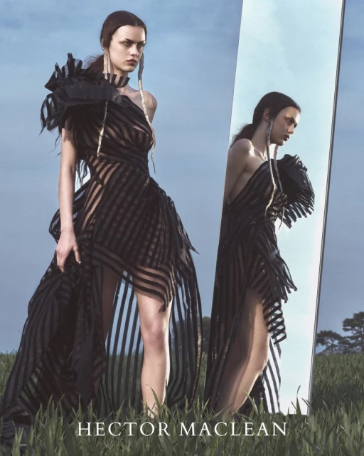 A fashion shot of a model in a long black striped dress with reflection in a standing mirror in a grassy field under a blue sky.