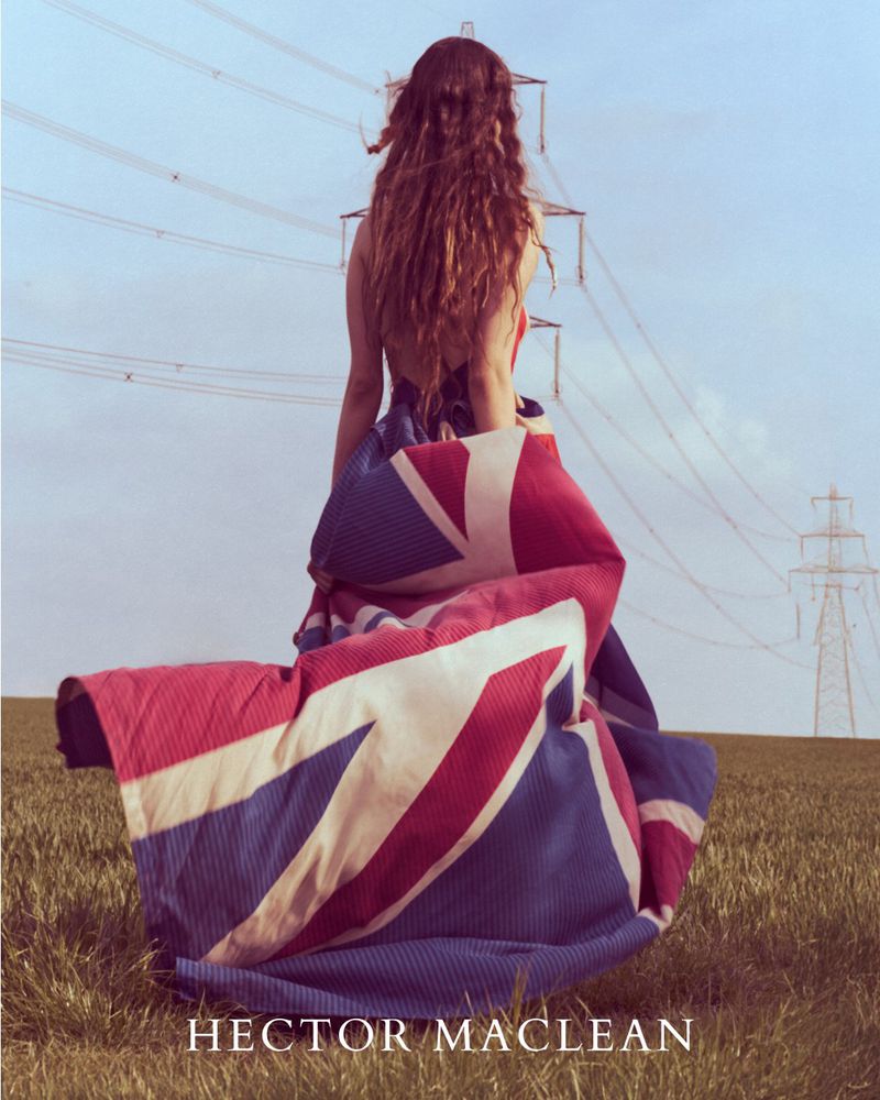 Woman stands in a field with powerlines in the background, draped in a Union Jack flag. Shot at eye-level on a partly sunny day.