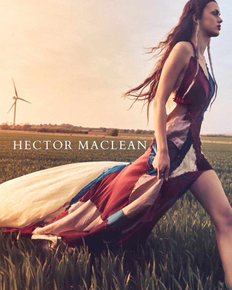 Woman in patterned dress walking in sunlit field with wind turbine in background for fashion editorial, outdoor fashion photography.