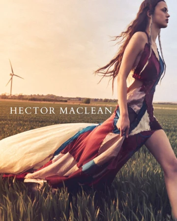 Woman in patterned dress walking in sunlit field with wind turbine in background for fashion editorial, outdoor fashion photography.