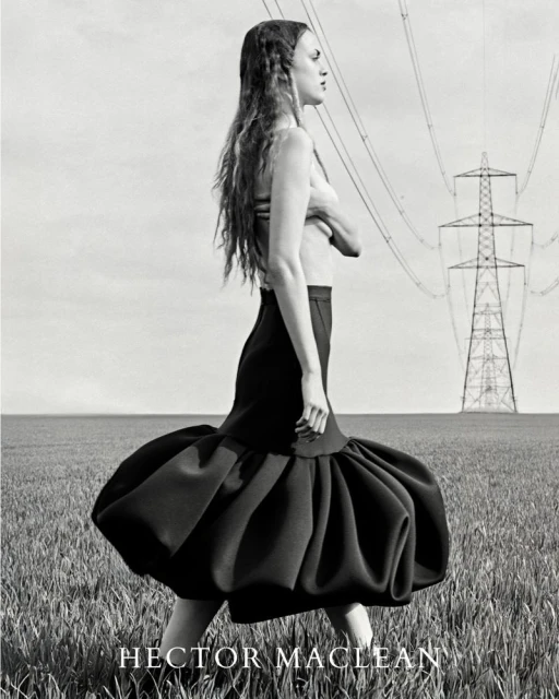 Artistic black and white fashion portrait of a woman in a dramatic skirt walking in a grassy field with an electrical tower in the background.