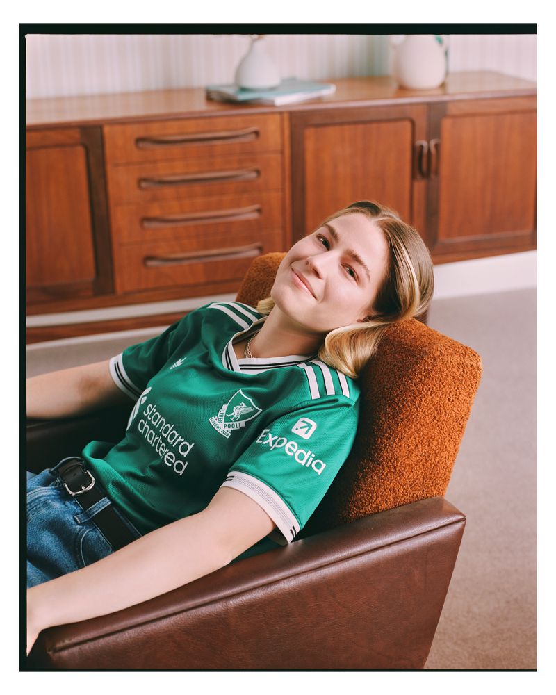 Young woman in a green soccer jersey lounges in a brown chair, set against a warm indoor backdrop featuring wooden furniture.