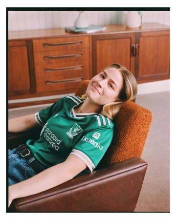 Young woman in a green soccer jersey lounges in a brown chair, set against a warm indoor backdrop featuring wooden furniture.